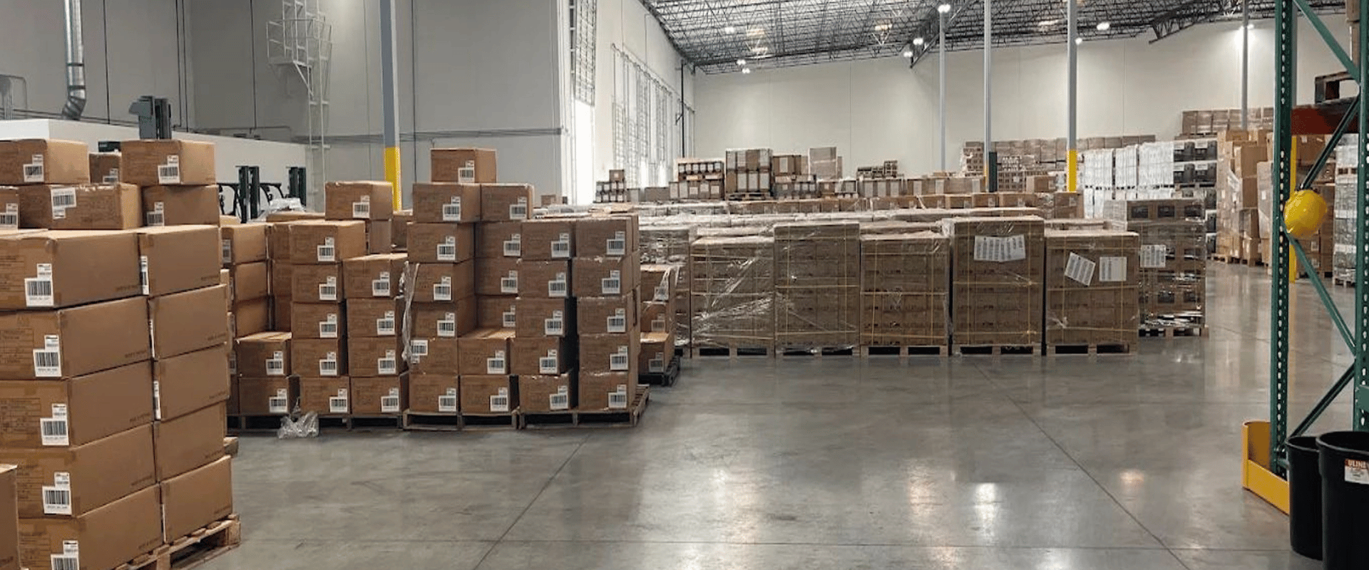 Hala logistyczna – palety z kartonami i towarem na magazynie, szerokie ciągi komunikacyjne w centrum dystrybucyjnym.