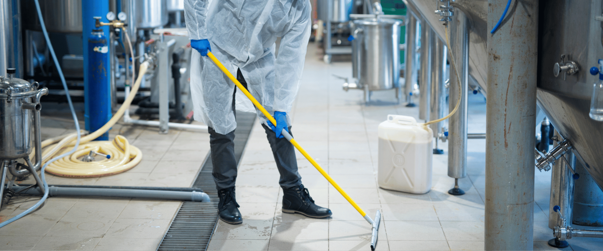Pracownik w odzieży ochronnej myje posadzkę w hali produkcyjnej, czyszcząc podłogę między stalowymi zbiornikami i instalacjami